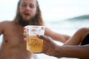 multiétnico grupo do amigos tendo Diversão em a praia, bebendo Cerveja e tendo Diversão foto