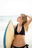 retrato do sorridente jovem mulher em pé com prancha de surfe às a de praia foto