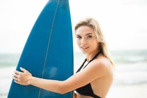 retrato do sorridente jovem mulher em pé com prancha de surfe às a de praia foto