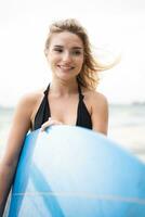 retrato do sorridente jovem mulher em pé com prancha de surfe às a de praia foto