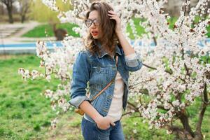 sexy mulher dentro jeans jaqueta, vermelho cheio lábios, ondulado Penteado posando perto flor árvore dentro Primavera dia. foto