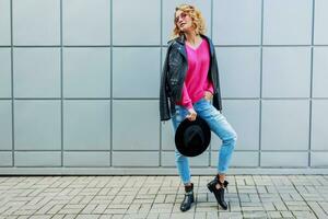 feliz loiro mulher posando em moderno ruas, bebendo café ou cappuccino. à moda outono roupa, couro Jaqueta e tricotado suéter. urbano fundo. Rosa oculos escuros. foto