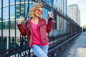 feliz loiro mulher posando em moderno ruas, bebendo café ou cappuccino. à moda outono roupa, couro Jaqueta e tricotado suéter. urbano fundo. Rosa oculos escuros. foto