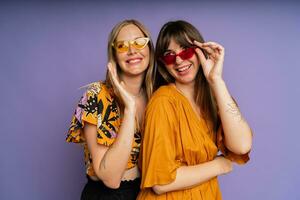 fechar acima retrato do dois à moda mulheres dentro oculos de sol e na moda verão roupas posando em roxa Bakground dentro estúdio. foto