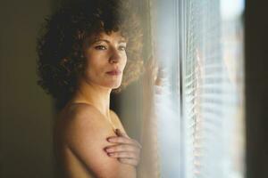 pensativo jovem nu mulher olhando ausente, inclinado em janela. foto