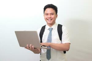 indonésio Senior Alto escola aluna vestindo branco camisa uniforme com cinzento gravata segurando uma computador portátil, em pé contra cinzento fundo foto