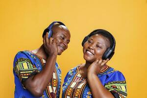 sorridente africano americano romântico casal vestindo étnico roupas desfrutando música juntos dentro sem fio fones de ouvido. homem e mulher com alegre expressão ouvindo para música dentro estúdio foto