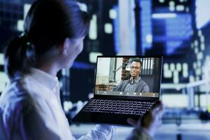 homem dentro teleconferência encontro com colega de trabalho enquanto passeando por aí cidade ruas às noite, dando ele trabalhos atualizações. cidadão usando computador portátil para mostrar colega período noturno metropolitano meio Ambiente foto