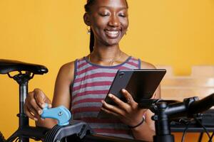 desportivo africano americano fêmea fixação bicicleta com Ferramentas e navegando digital dispositivo para soluções. detalhado tiro do saudável Preto mulher procurando em telefone tábua para bicicleta manutenção. foto