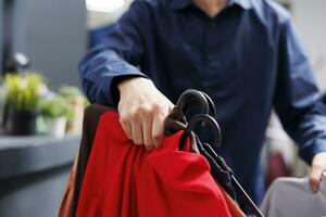 fechar acima do masculino mão segurando roupas em cabides enquanto compras dentro roupas loja, seletivo foco. homem cliente escolhendo vestuário às moda tomada, olhando Apesar vestuário prateleiras com roupas Itens foto