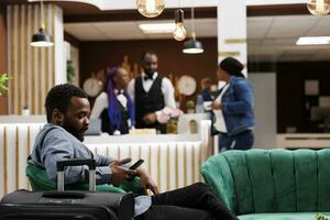 africano americano cara viajante sentado dentro hotel lobby com Smartphone conversando conectados com amigos enquanto esperando para hotel check-in, usando livre Wi-fi. homem turista com bagagem esperar às recepção área foto