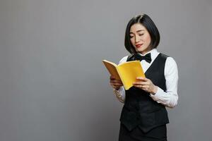 pensativo ásia garçonete vestido dentro profissional uniforme lendo livro com amarelo cobrir. jovem atraente mulher recepcionista segurando capa mole Diário enquanto posando dentro estúdio foto