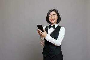 sorridente ásia garçonete usando Smartphone e olhando às Câmera com amigáveis facial expressão. alegre restaurante recepcionista dentro profissional uniforme segurando Móvel telefone retrato foto