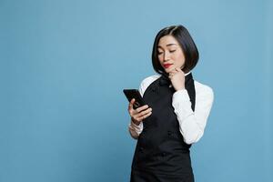 jovem ásia garçonete digitando em Smartphone, verificação conectados ordens para restaurante. hotel atraente mulher recepcionista dentro uniforme mensagens de texto em Móvel telefone, respondendo cliente mensagem foto