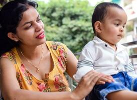 amoroso mãe carregando do dela bebê às casa sacada. brilhante retrato do feliz mãe segurando criança dentro dela mãos. mãe com dela pequeno 1 ano velho filho. foto