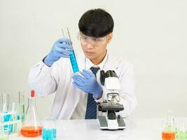 retrato ásia homem aluna cientista vestindo uma médico vestido dentro a laboratório olhando mão às químico. causou de mistura reagentes dentro científico pesquisa laboratórios com teste tubos e microscópio em a mesa foto