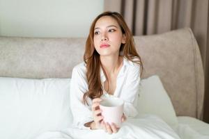 retrato linda mulher asiática acordar e segurando uma xícara de café ou caneca na cama pela manhã foto