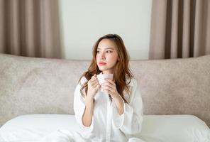 retrato linda mulher asiática acordar e segurando uma xícara de café ou caneca na cama pela manhã foto