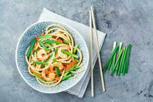 udon Macarrão e wok com camarões, pimentas e cebolas dentro uma prato em uma guardanapo e pauzinhos em a mesa. chinês Comida. topo Visão foto