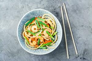 udon e wok Macarrão com camarões, pimentas e cebolas dentro uma prato e pauzinhos em a mesa. chinês Comida. topo Visão foto