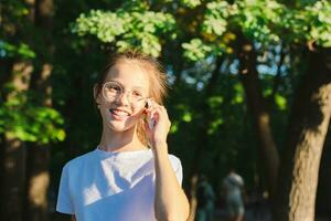 uma menina dentro óculos alegremente fala em uma botão de apertar Telefone entre a árvores foto