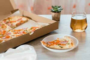 tirar Comida. uma fatia do pizza dentro uma descartável plástico placa, Cerveja e uma caixa do pizza em a mesa dentro a cozinha. foto