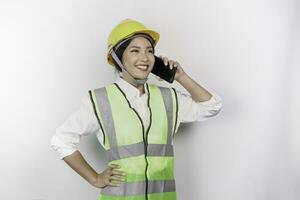 uma sorridente ásia mulher trabalho vestindo segurança capacete e colete enquanto tendo uma telefone chamar, isolado de branco fundo. trabalho dia conceito. foto