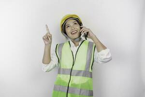 uma sorridente ásia mulher trabalho vestindo segurança capacete e colete, falando em dela telefone e apontando para cópia de espaço acima dela, isolado de branco fundo. trabalho dia conceito. foto