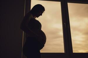 grávida mulher em pé ao lado janela e segurando dela barriga. silhueta do grávida mulher. foto