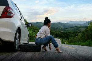 jovem mulher asiática sentada perto do carro esperando por ajuda foto