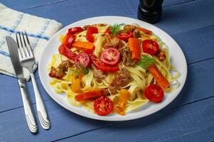 fettuccine com grelhado carne, cenouras, Sino pimentas, aneto tomates em uma azul de madeira fundo. massa com legumes. lado visualizar. foto