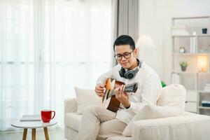 ásia homem vestindo óculos e fone de ouvido jogando guitarra enquanto sentado em sofá dentro a vivo quarto às lar. ásia homem escrevendo música enquanto jogando guitarra às lar. compor música música conceito. foto