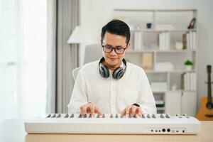 homem asiático usando fones de ouvido brancos toca piano e canta uma música e aprende on-line com o celular e compõe a música ou grava o som com um microfone e usa o programa de gravação de música do computador foto