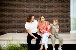 família com uma mãe, pai e filha sentado lado de fora em a passos do uma frente varanda do uma tijolo casa foto