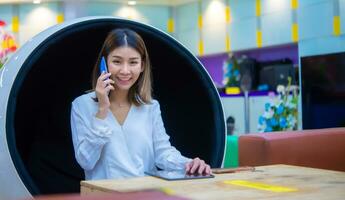 lindo ásia o negócio mulher sentado em moderno volta cadeira e usando telefone para comunicar com cliente com sorrir enquanto olhando para a Câmera, digital marketing. foto