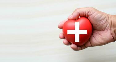 uma vermelho Cruz é uma símbolo do médico saúde. mão segurando uma coração e em uma de madeira escrivaninha. cópia de espaço para a texto e conteúdo. vermelho Cruz RCP, mundo coração dia, mundo saúde dia, sangue órgão doação foto