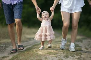 feliz um ano de idade menina é ser mantido de Papai e mãe. pequeno criança caminhando com pais. uma fofa bebê aprende para andar com a Socorro do dela pais. foto