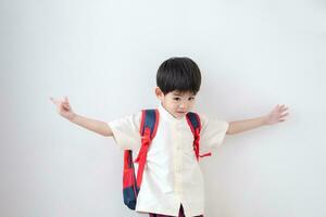 ásia Garoto vestindo tradicional tailandês roupas, em pé com uma escola bolsa. faço uma acenando gesto preparando para ir para escola em uma branco fundo foto