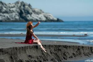 mulher dentro vermelho grandes vestir com elevado bainha sentado em arenoso de praia com nu pernas pendurado sobre penhasco foto