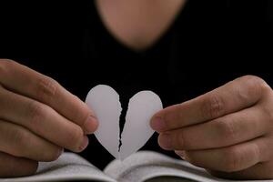 fêmea mão segurando uma meio rasgado branco papel coração sobre a aberto livro em a mesa. branco papel coração rasgado dentro metade dentro mão foto