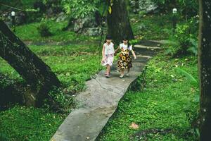 dois fofa irmãs caminhando em uma pedra caminho dentro uma botânico jardim com verde plantas e colorida flores em volta. crianças estudando natureza foto