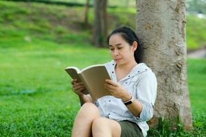 alegre jovem mulher dentro Jaqueta lendo uma livro dentro verão parque. concentrado mulher sentado em Relva e estudando debaixo árvore durante período de férias. Educação conceito foto