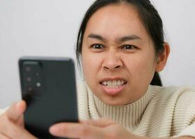 retrato do ásia mulher segurando Móvel telefone, lendo mau mensagem, sentindo-me insatisfeito e nervoso. chateado jovem mulher sentado às casa segurando uma Smartphone dentro dela mão recebe uma mensagem com mau notícias. foto