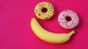 dois donuts e uma banana repousam na superfície rosa, formando uma emoção de sorriso
