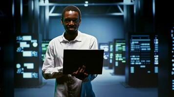 retrato do sorridente admin usando computador portátil para avaliar energia consumo através servidor montagens em rack componentes. profissional fazer certo dados Centro mainframes temperatura sensores estão corrida suavemente foto