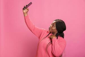linda africano americano mulher fazendo selfie com Móvel telefone enquanto posando dentro estúdio sobre Rosa fundo. à moda alegre menina levando fotografia desfrutando postagem em social meios de comunicação foto