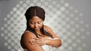 africano americano menina sentindo-me radiante aplicando creme dentro estúdio, promovendo cuidados com a pele produtos para cosméticos campanha. sem falhas mulher usando hidratante ou sérum em Câmera, auto amar. foto