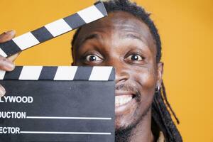 retrato do homem segurando quadro-negro para cortar cenas dentro filme indústria, posando em amarelo fundo. africano americano jovem adulto trabalhando dentro filme fazer Produção e cinematografia foto