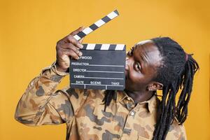 retrato do sorridente africano americano homem segurando filme Produção quadro-negro em pé dentro estúdio sobre amarelo fundo. alegre pessoa dando audição para entretenimento vídeo. cinema conceito foto