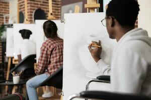 artes Atividades para adultos com fisica deficiências. Desativado africano americano homem cadeira de rodas do utilizador desenhando com lápis em tela de pintura enquanto visitando grupo arte aula, atendendo terapêutico sessão foto